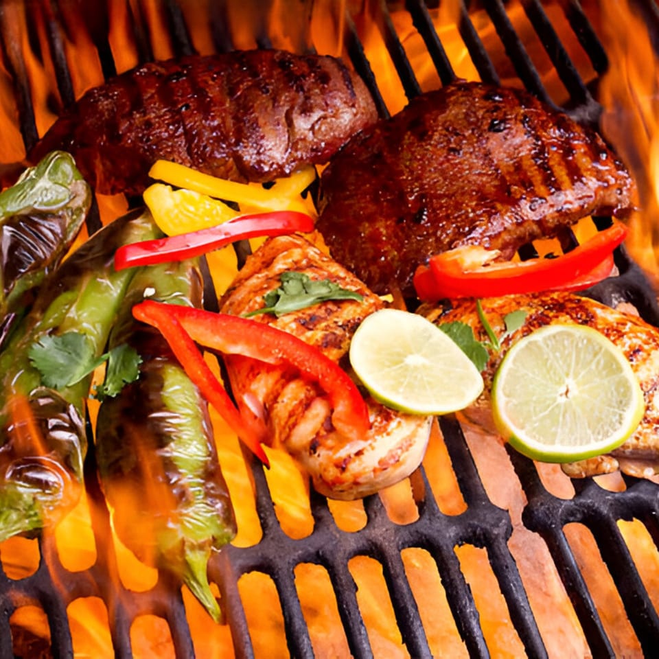 Mexican grilled steak, chicken, peppers, and limes cooking over open flames on a grill.