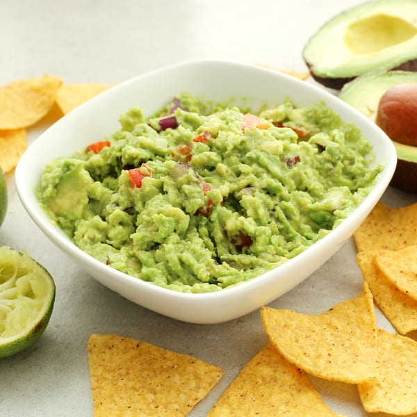 Fresh guacamole in a white bowl, surrounded by tortilla chips, lime, and avocado halves.