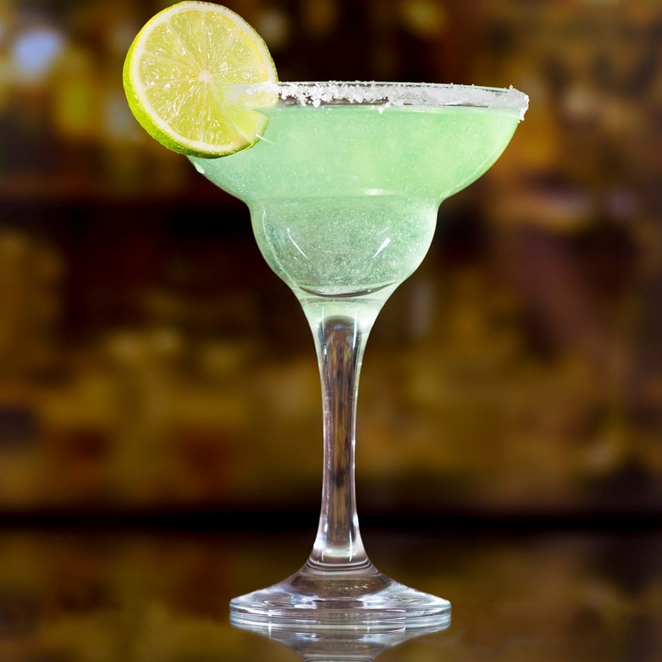 A vibrant green La Parrilla margarita in a salt-rimmed glass, garnished with a fresh lime slice.