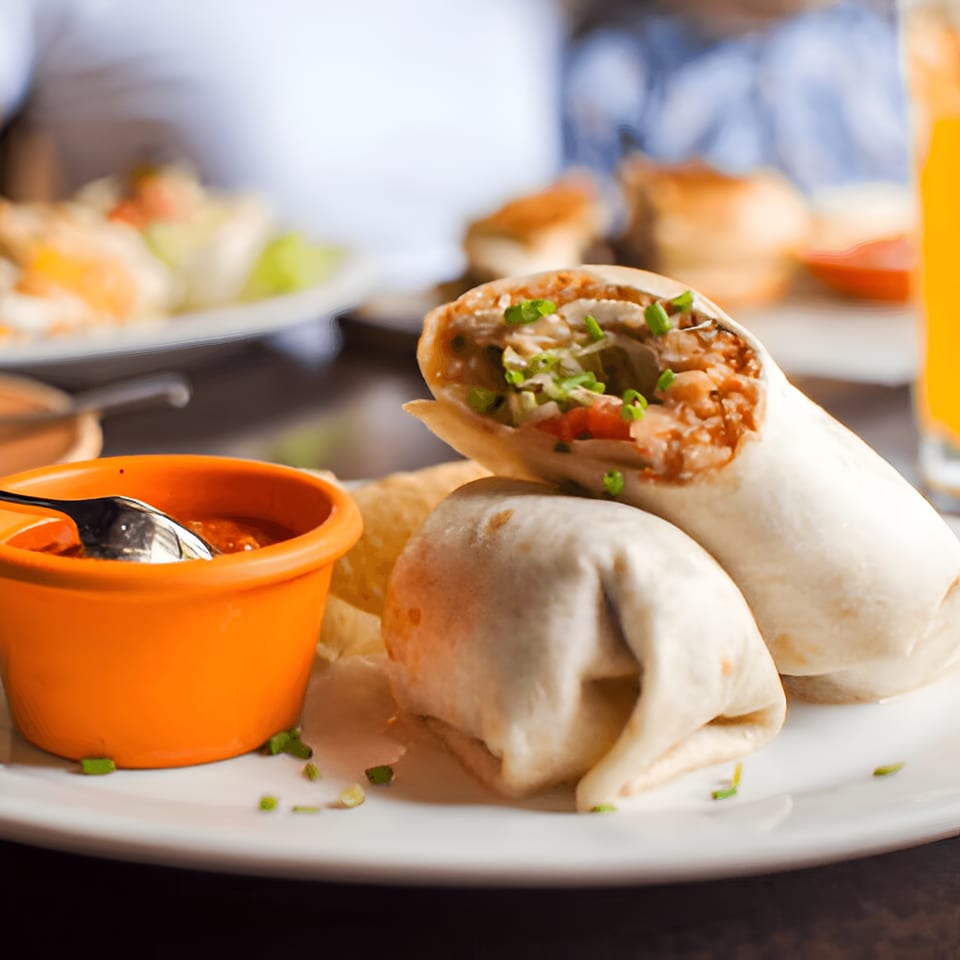 Two La Parrilla burritos with an orange bowl of salsa and chips on a white plate.